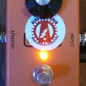 Puede incluir: Pedal de efectos de guitarra de color naranja con una perilla negra, una pegatina blanca con el texto "Alchemy Audio" y un rayo rojo, y el texto "phase 90".
