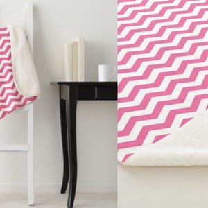 May include: A pink and white chevron pattern blanket with a white sherpa lining. The blanket is draped over a white ladder and displayed on a white surface. A black table and white mug are in the background.