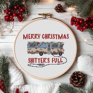 May include: A white embroidery hoop with a red and white cross-stitch design. The design features a cartoon RV with Christmas lights and the text "Merry Christmas Shitter's Full".