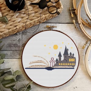 May include: A cross-stitch embroidery hoop with a white background and a design of Hogwarts castle emerging from a book. The castle is gray with yellow windows and a red roof. The book is open with pages that are beige and brown. There are yellow stars and a yellow sun in the sky above the castle.