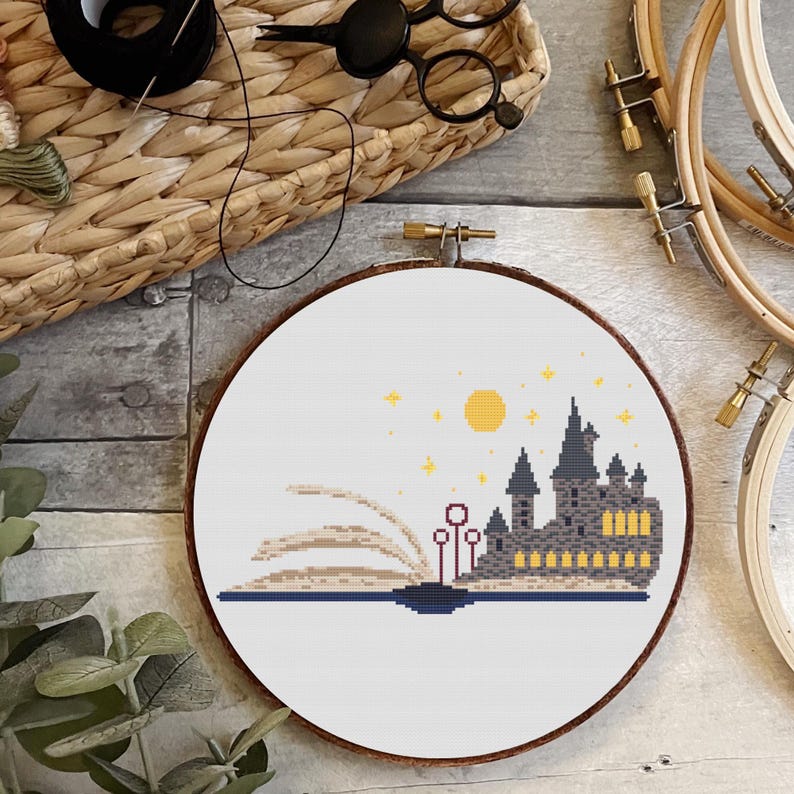 May include: A cross-stitch embroidery hoop with a white background and a design of Hogwarts castle emerging from a book. The castle is gray with yellow windows and a red roof. The book is open with pages that are beige and brown. There are yellow stars and a yellow sun in the sky above the castle.