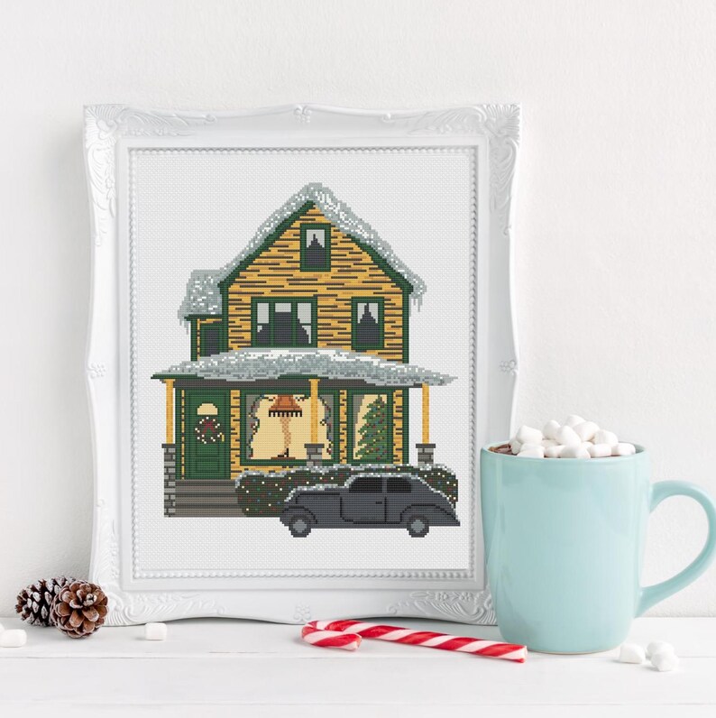 May include: A cross-stitch pattern of a yellow house with a green roof and white trim. The house is covered in snow and has a black car parked in front of it. The house has a porch with a wreath on the door and a Christmas tree in the window.