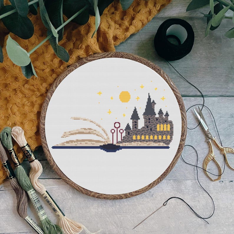May include: A white cross stitch embroidery hoop with a design of Hogwarts castle emerging from an open book. The book is surrounded by stars and a yellow moon. The design is on white Aida cloth.