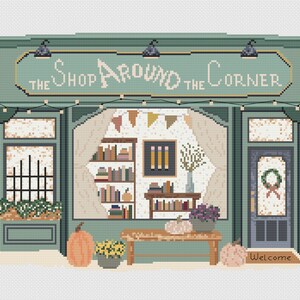 May include: A cross-stitch design of a shop facade with the text "The Shop Around the Corner". The shop is painted in shades of green and features a window display with books, pumpkins, and flowers. A welcome mat is at the entrance.