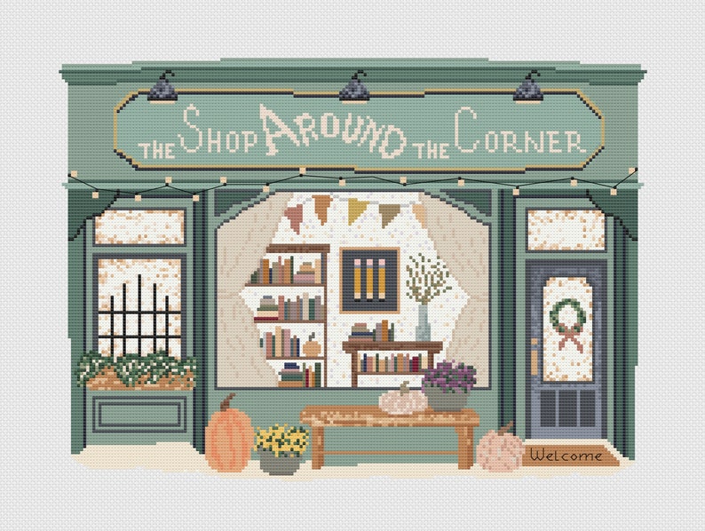 May include: A cross-stitch design of a shop facade with the text "The Shop Around the Corner". The shop is painted in shades of green and features a window display with books, pumpkins, and flowers. A welcome mat is at the entrance.