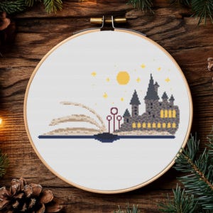May include: A white cross-stitch embroidery hoop with a design of Hogwarts castle emerging from an open book. The castle is gray with yellow windows and a red roof. The book is beige with a brown spine. The background is white with a yellow moon and stars.
