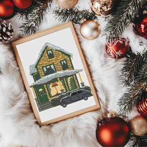 May include: A framed cross-stitch pattern of a yellow house with green trim and a black car in front of it. The house is covered in snow and has a Christmas tree in the window.