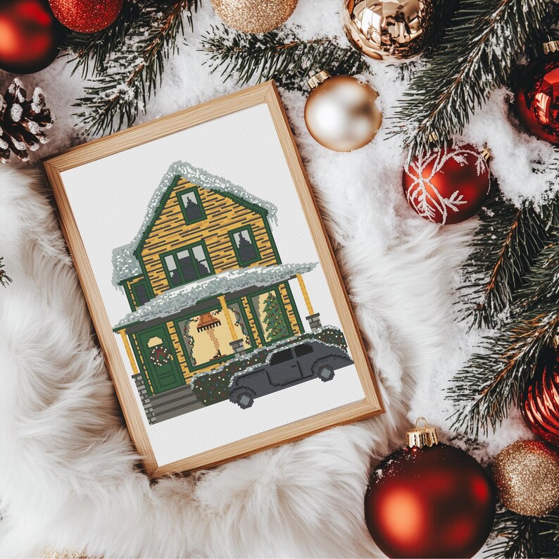 May include: A framed cross-stitch pattern of a yellow house with green trim and a black car in front of it. The house is covered in snow and has a Christmas tree in the window.