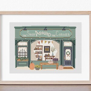 May include: A framed cross-stitch of a shop facade with the text "The Shop Around the Corner." The shop has a green awning, string lights, and a window display with books, pumpkins, and flowers. A welcome mat is at the door.