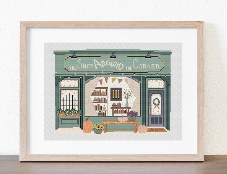 May include: A framed cross-stitch of a shop facade with the text "The Shop Around the Corner." The shop has a green awning, string lights, and a window display with books, pumpkins, and flowers. A welcome mat is at the door.