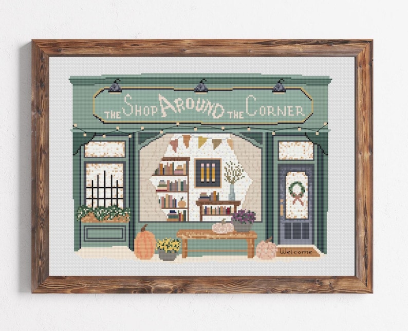 May include: Cross-stitch art of a shop facade with the text "The Shop Around the Corner." The shop is painted in shades of green and features pumpkins, flowers, and a welcome mat. The artwork is framed in a brown wooden frame.