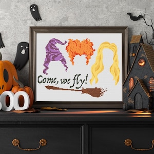 Pode incluir: Padrão de ponto cruz com três bruxas com o texto "Come, we fly!" e uma vassoura. As bruxas têm cabelo roxo, laranja e loiro. O padrão é emoldurado e exibido em uma prateleira com decorações de Halloween.