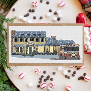 May include: A cross-stitch pattern of a yellow house with a red and green bus in front of it. The house has a white roof and is decorated with Christmas lights. The bus is decorated with a wreath and Christmas lights.