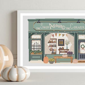 May include: A framed cross-stitch art piece depicts a shop with the text "The Shop Around the Corner". The shop is green with a welcoming door and a display of books. Two decorative pumpkins sit in front of the frame.
