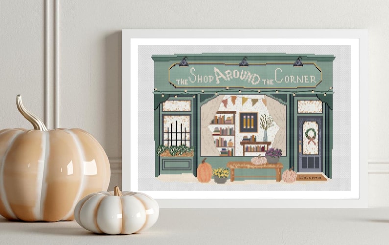 May include: A framed cross-stitch art piece depicts a shop with the text "The Shop Around the Corner". The shop is green with a welcoming door and a display of books. Two decorative pumpkins sit in front of the frame.