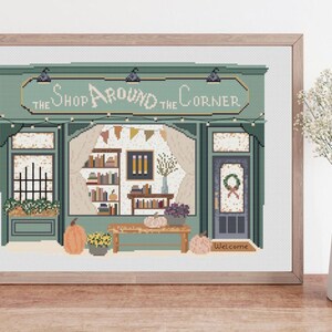 May include: Cross-stitch art of a shop facade with the text "The Shop Around the Corner." The shop is painted in shades of green and features a door, windows, and a welcoming mat. Pumpkins and flowers decorate the front.