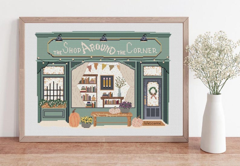 May include: Cross-stitch art of a shop facade with the text "The Shop Around the Corner." The shop is painted in shades of green and features a door, windows, and a welcoming mat. Pumpkins and flowers decorate the front.