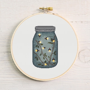 May include: A cross-stitch embroidery of a mason jar filled with fireflies. The jar is a muted teal color with a silver lid. Inside, yellow and black fireflies glow among dark branches. The embroidery is set in a wooden hoop.