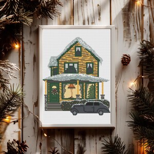 May include: A cross-stitch pattern of a yellow house with a white roof and snow on the roof. The house has a green door and a porch with a black car parked in front. There is a Christmas tree in the window and a wreath on the door.