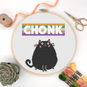 May include: A cross-stitch embroidery hoop with a black cat and the word "CHONK" in rainbow letters. The cat is sitting with its paws tucked under its body and has pink cheeks.