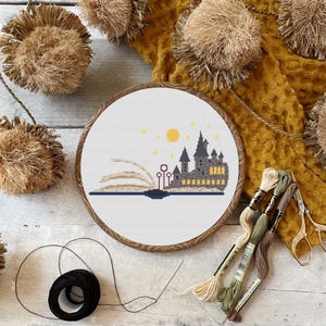 May include: A cross-stitch embroidery hoop with a white background and a design of Hogwarts castle emerging from a book. The castle is brown and gray with yellow windows. The book is open and has a brown cover. There are stars and a yellow moon in the sky above the castle.