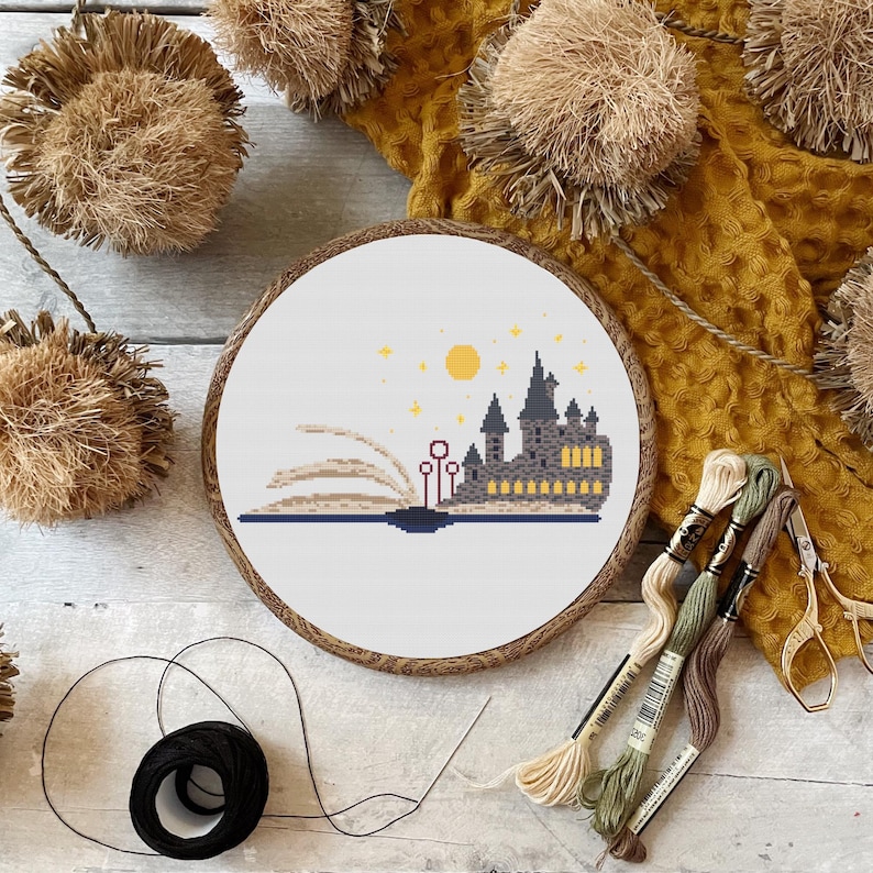 May include: A cross-stitch embroidery hoop with a white background and a design of Hogwarts castle emerging from a book. The castle is brown and gray with yellow windows. The book is open and has a brown cover. There are stars and a yellow moon in the sky above the castle.