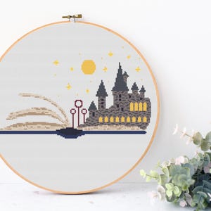 May include: A cross-stitch pattern of Hogwarts castle, a magical school from the Harry Potter series, emerging from an open book. The castle is depicted in gray and black with yellow windows, and the book is beige with a brown spine. The background is white with a yellow moon and stars.