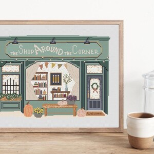 May include: Cross-stitch art of a shop facade with the text "The Shop Around the Corner." The shop is painted in shades of green and features a door, windows, and a sign. Pumpkins and flowers decorate the scene, creating a cozy, autumnal feel.
