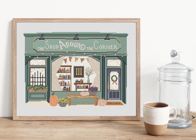 May include: Cross-stitch art of a shop facade with the text "The Shop Around the Corner." The shop is painted in shades of green and features a door, windows, and a sign. Pumpkins and flowers decorate the scene, creating a cozy, autumnal feel.