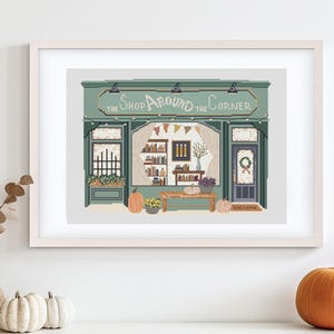 May include: A framed print of a shop with the text "The Shop Around the Corner." The shop is painted in shades of green and has a welcoming autumn theme, with pumpkins and floral arrangements.