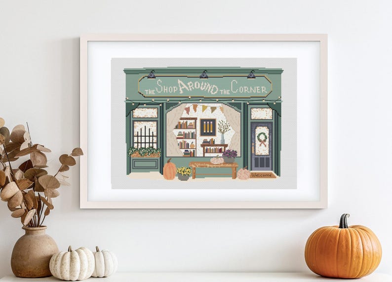 May include: A framed print of a shop with the text "The Shop Around the Corner." The shop is painted in shades of green and has a welcoming autumn theme, with pumpkins and floral arrangements.