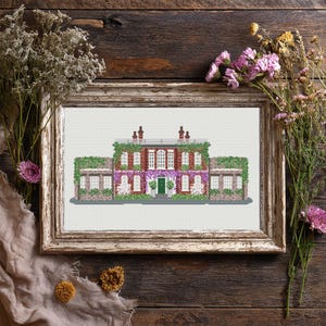Puede incluir: Arte de punto de cruz de una casa señorial de ladrillo con puerta verde, rodeada de enredaderas y flores. La obra enmarcada se exhibe sobre una superficie de madera rústica, con flores secas y detalles de tela.