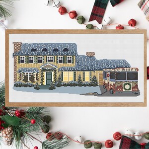 May include: A cross-stitch pattern of a yellow house with a blue roof and a snow-covered lawn. A red and white Christmas bus is parked in front of the house.