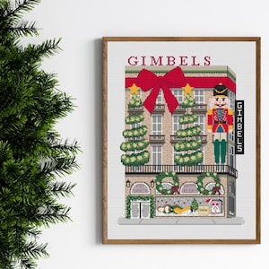 Peut inclure: Une broderie au point de croix encadrée représentant un bâtiment décoré pour Noël avec le mot "GIMBELS" en haut. Elle présente un grand nœud rouge, des sapins et un casse-noisette. La devanture affiche "Visit Santa" et "Happy Holidays".