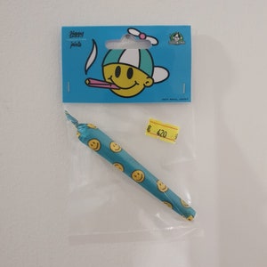 以下が含まれることがあります： スマイリーフェイスが描かれた青と黄色のパッケージ。パッケージには、スマイリーフェイスのパターンが描かれた偽のジョイントが入っています。価格タグには「£ 420」と表示されています。