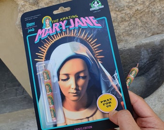 Peut inclure: Une boîte colorée avec le texte "The Amazing Mary Jane Saints Edition" et une image d'une femme en robe bleue. La boîte a un autocollant jaune qui dit "Priez pour nous".