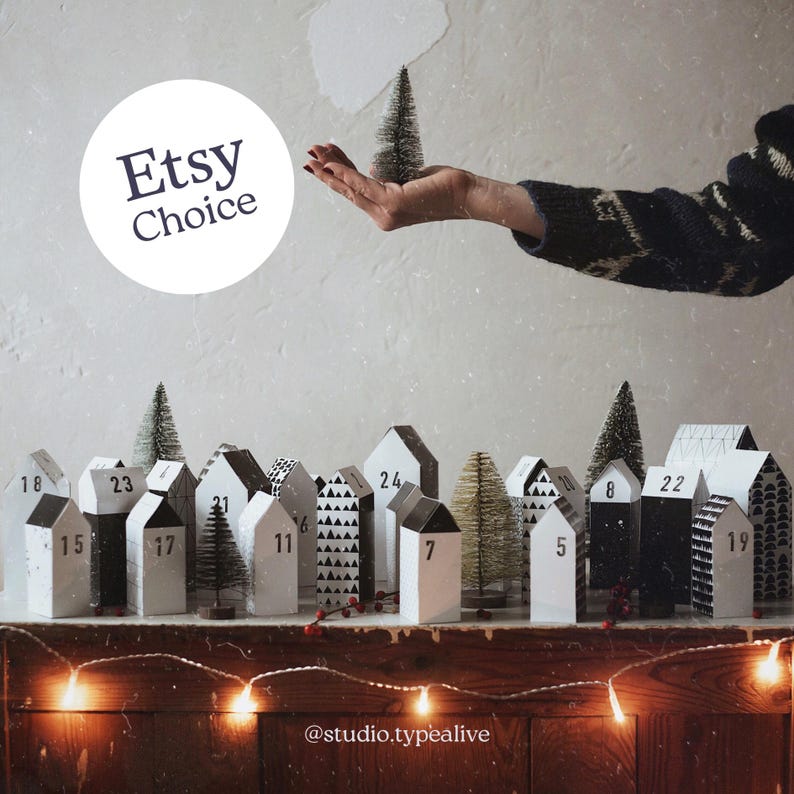 May include: A black and white paper advent calendar with numbered houses and small trees. The calendar is on a wooden surface with a string of lights. The text "Etsy Choice" is in a circle above the calendar.