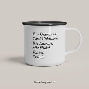 Könnte beinhalten: Weißer Emaillebecher mit schwarzem Rand und Henkel. Der Becher hat schwarzen Text in einer einzigartigen Schriftart. Der Text lautet: "Ein Glühwein. Swei Glühweih. Rei Lühwei. Hie Hühei. Flünei. Snlwln." Der Becher hat den Instagram-Handle @studio.typealive.