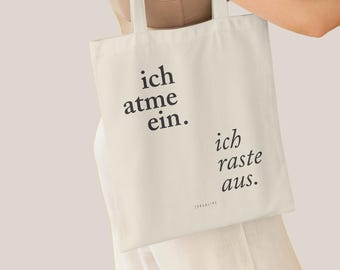 May include: A white canvas tote bag with black text that reads "ich atme ein. ich raste aus." The text is centered on the bag. The bag is being held by a person.