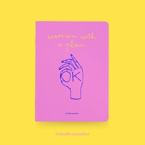 May include: A pink notebook with the text "woman with a plan" in yellow script. A blue outline of a hand making the "OK" gesture is below the text. The notebook has the text "@studio.typealive" at the bottom.