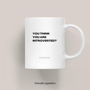 Peut inclure: Mug en céramique blanche avec une anse incurvée. Le mug affiche le texte "YOU THINK YOU ARE INTROVERTED?" en noir, avec "TYPEALIVE" imprimé en dessous. Le mug est de conception simple mais élégante.