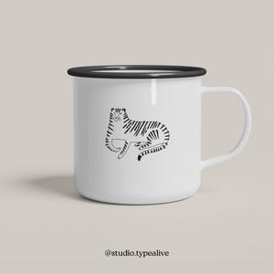 Könnte beinhalten: Eine weiße Emailletasse mit schwarzem Rand und Henkel. Auf der Vorderseite der Tasse ist eine Schwarzweiß-Zeichnung eines Tigers aufgedruckt. Der Text "@studio.typealive" ist unter dem Bild aufgedruckt.