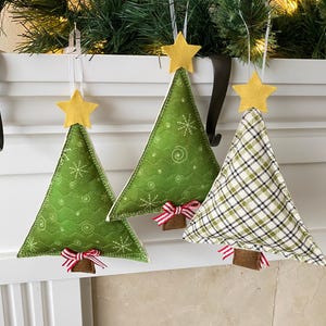 May include: Three felt Christmas tree ornaments hanging on a white mantel. The ornaments are green with a yellow star on top and a red and white ribbon at the bottom. One ornament has a plaid pattern.