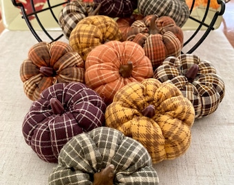 Puede incluir: Una colección de calabazas de tela en varios colores y patrones, incluyendo naranja, amarillo y diseños de cuadros. Las calabazas están dispuestas sobre una superficie neutra, con algunas dentro de una cesta de alambre negro, evocando un tema de cosecha otoñal.
