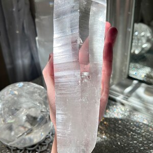May include: A large, clear quartz crystal with a pointed tip and a smooth, polished surface. The crystal is held in a hand, and the background is blurred.