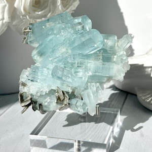 May include: A cluster of pale blue aquamarine crystals, mounted on a clear acrylic stand. The crystals have a translucent quality, with visible facets and a layered structure. The base of the crystal cluster has dark grey mineral inclusions.