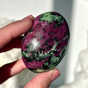 May include: A smooth, oval gemstone with a blend of colours. The stone displays a deep magenta hue with patches of green and black. The gemstone is held between fingers, highlighting its smooth, rounded form.