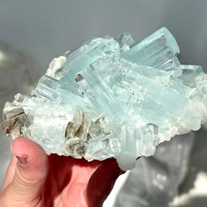 May include: A cluster of aquamarine crystals, showcasing a pale blue hue. The crystals are elongated and translucent, with some areas appearing opaque. The mineral formation includes small, gray-brown mica flakes.