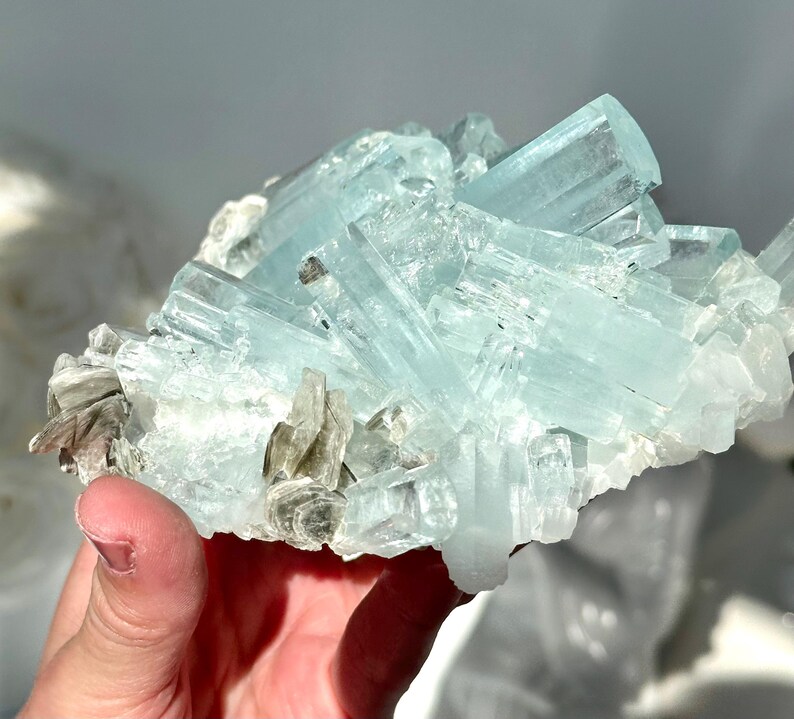 May include: A cluster of aquamarine crystals, showcasing a pale blue hue. The crystals are elongated and translucent, with some areas appearing opaque. The mineral formation includes small, gray-brown mica flakes.