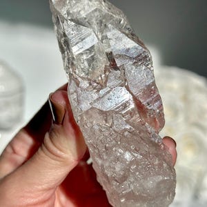 May include: A clear quartz crystal with a rough, textured surface. The crystal is held in a hand and has a natural, irregular shape. The quartz has a translucent appearance with light reflecting off its facets.